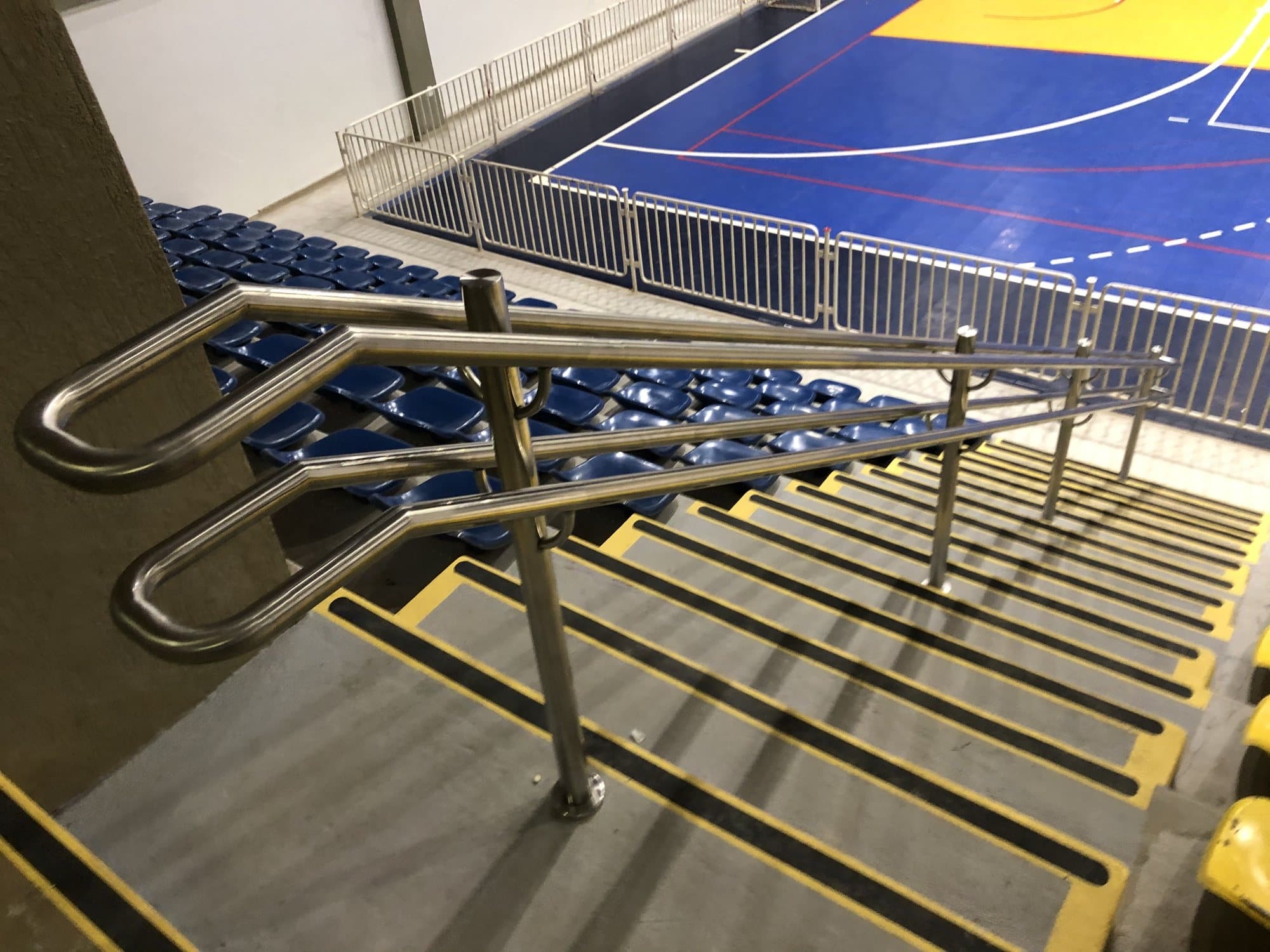 Início do corrimão duplo em aço inox no topo da escada da arquibancada, com a quadra ao fundo