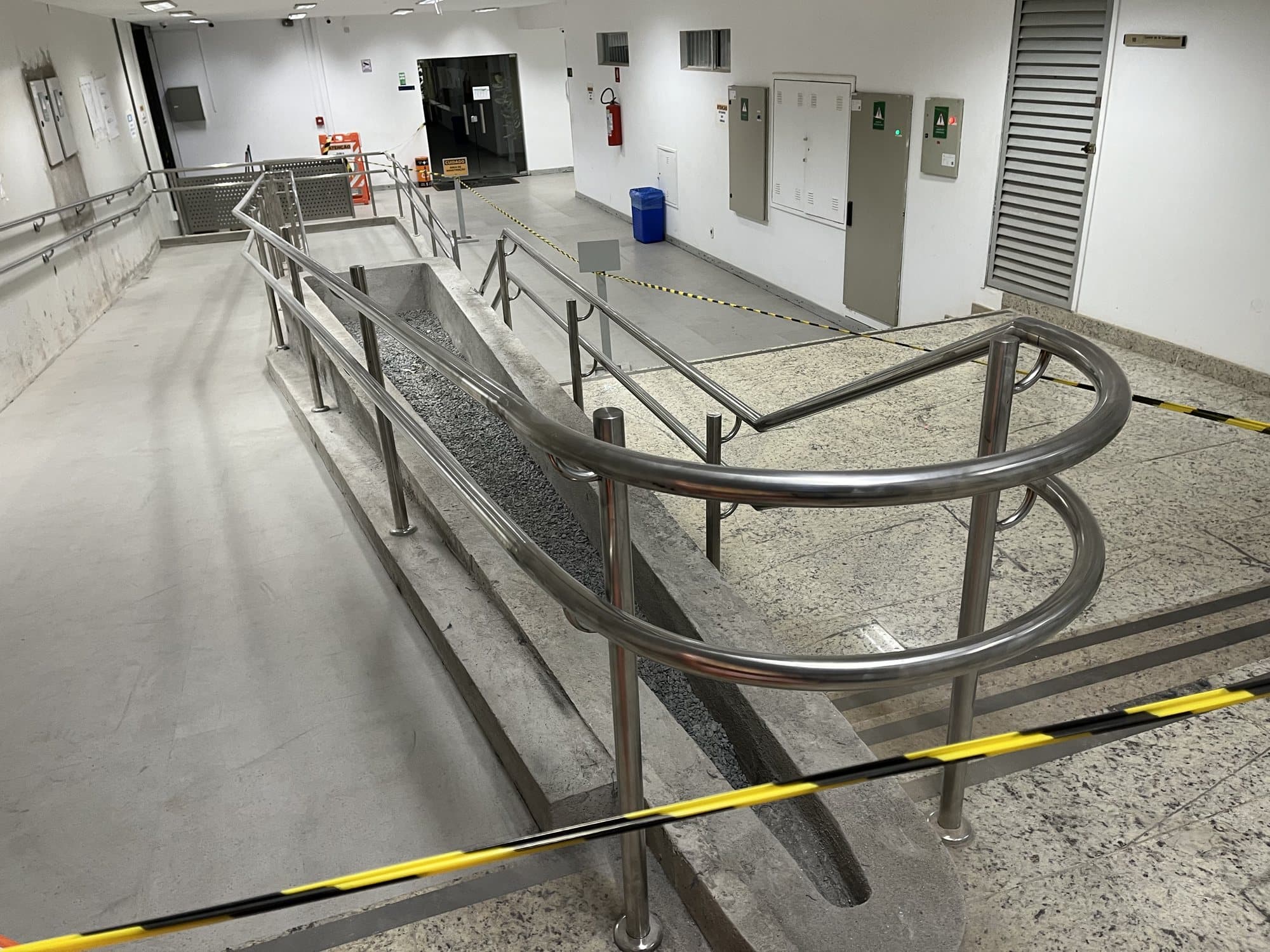 Corrimão duplo em aço inox com arremate curvo no início de uma rampa de acessibilidade em edifício