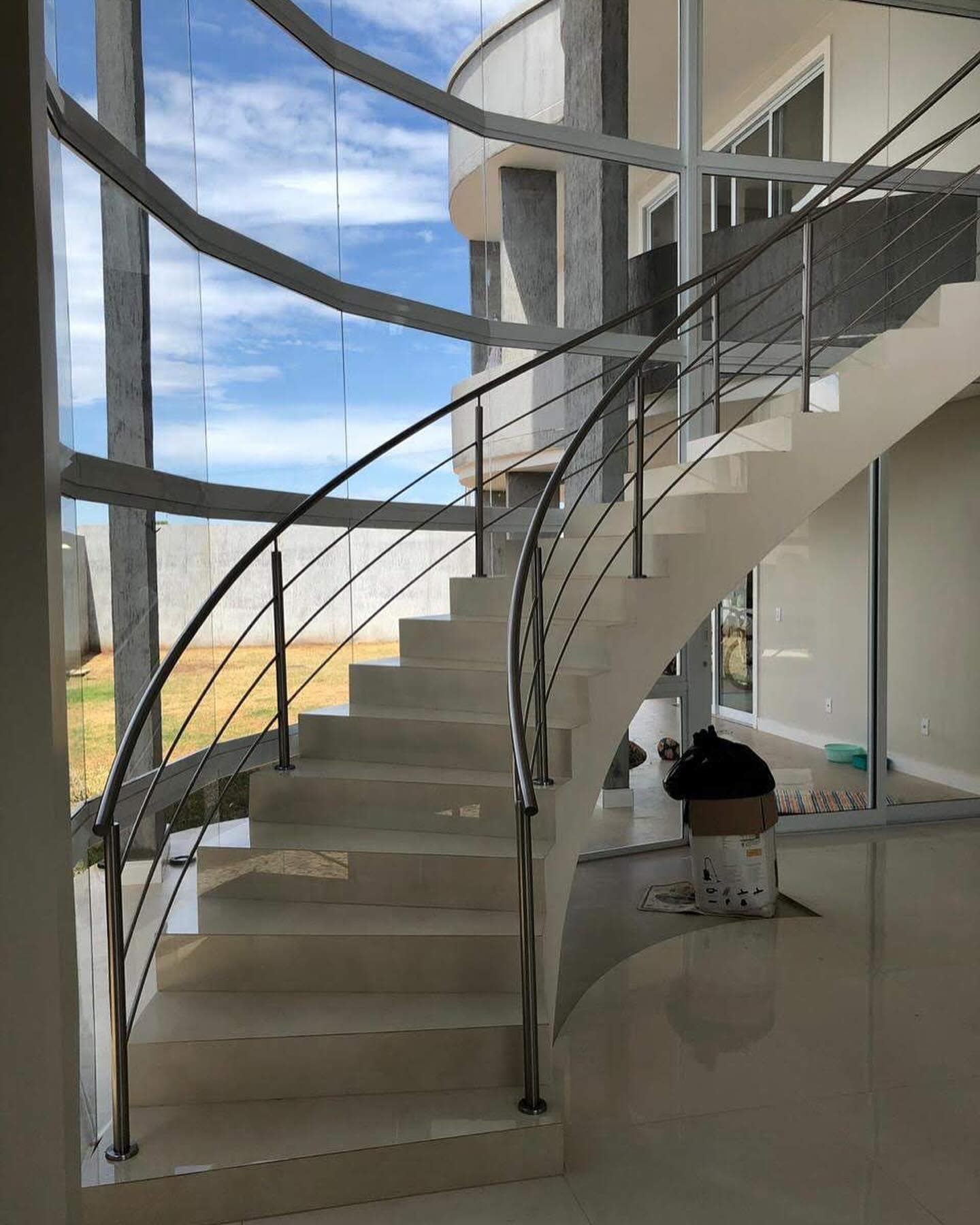Escada curva de degraus claros com corrimão e guarda-corpo em aço inox, diante de uma fachada de vidro