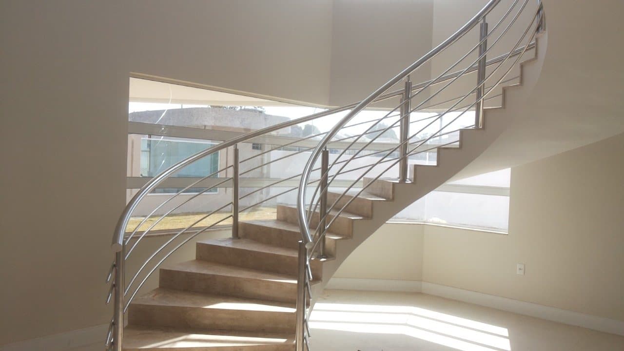 Escada curva em mármore com guarda-corpo em aço inox de tubos curvados, diante de uma janela