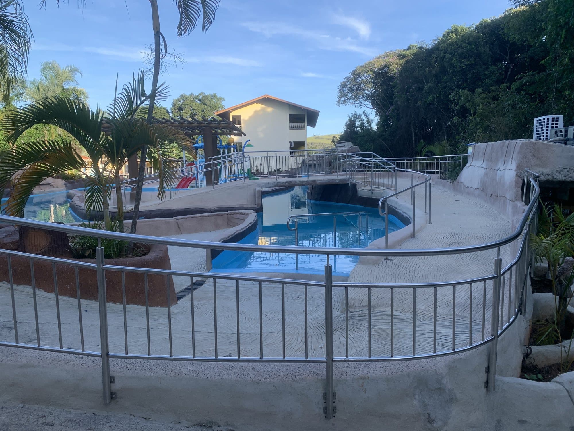 Guarda-corpo em aço inox contornando a rampa curva de acesso a uma piscina, com palmeiras ao fundo