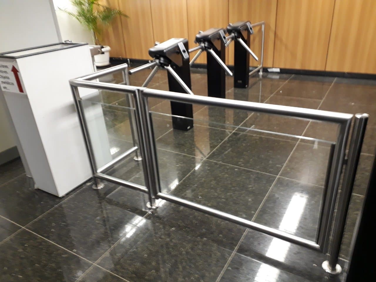 Guarda-corpo em aço inox com vidro ao lado das catracas de uma recepção com piso de granito escuro