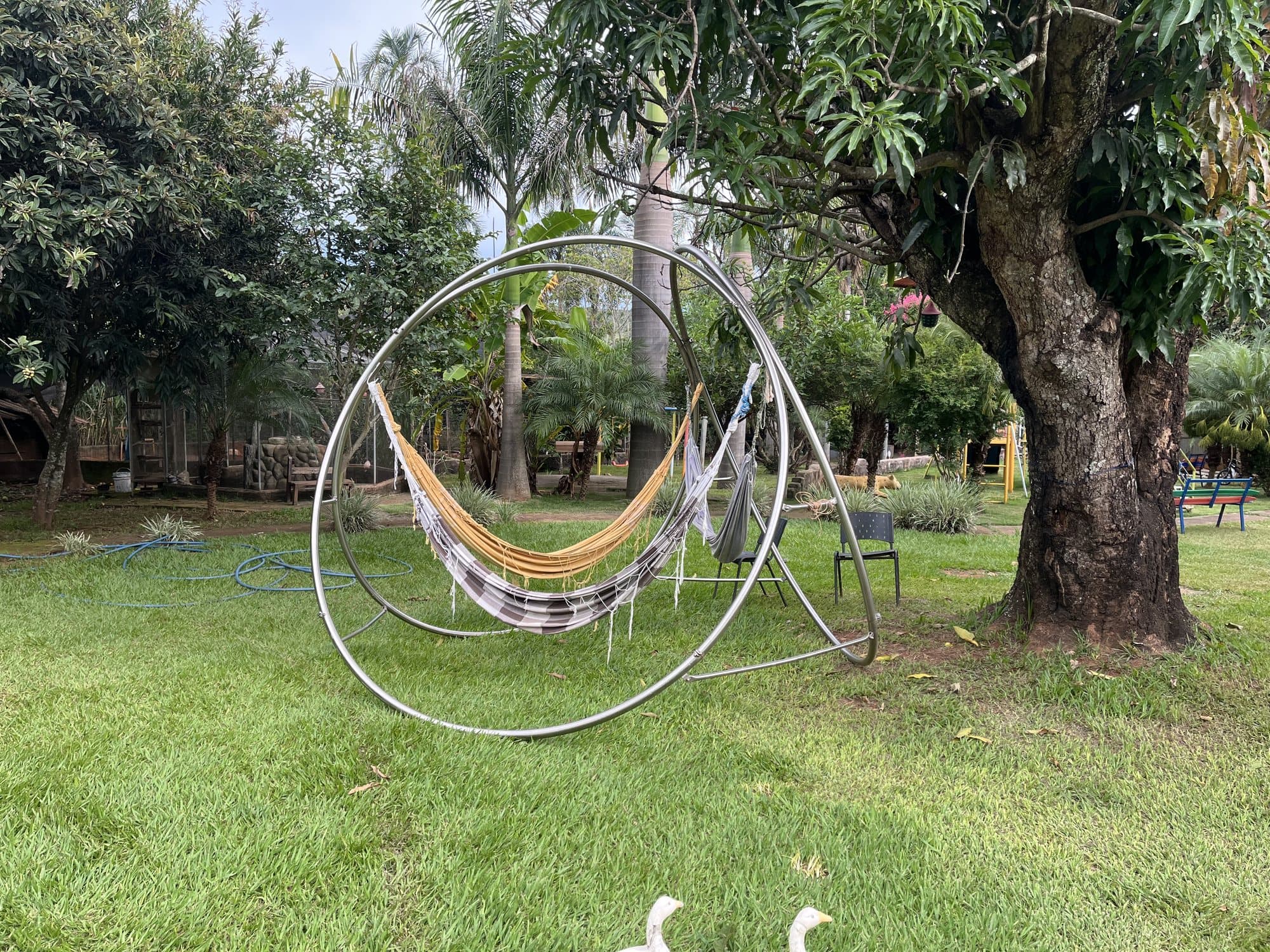 Suporte em aço inox de aros cruzados com redes de descanso, visto de lado em um jardim